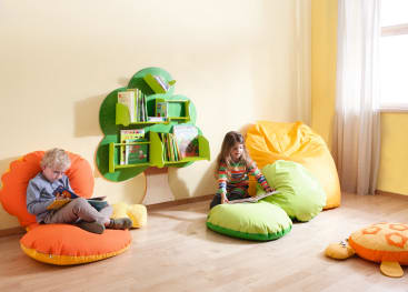Zwei kleine Kinder sitzen auf hellen Sitzsäcken und lesen in einem sonnigen Zimmer Bücher. Ein baumförmiges Bücherregal mit mehreren Büchern ist an der Wand zwischen ihnen angebracht. Licht strömt durch ein Fenster mit weißen Vorhängen herein.