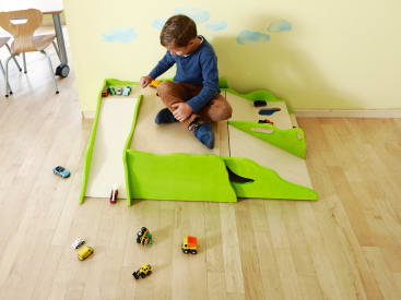 Ein kleiner Junge sitzt auf einem Holzboden und spielt mit Spielzeugautos und -lastwagen auf einem grün-beigen Holzspielzeug in Form von Rampen und Straßen. Mehrere Spielzeugfahrzeuge sind um ihn herum verstreut. An die Wand dahinter sind Wolken gemalt.