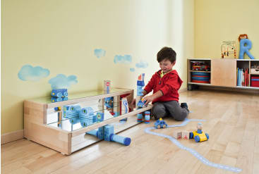 Ein kleiner Junge mit rotem Kapuzenpulli sitzt auf einem Holzboden und spielt mit blauen Bauklötzen und Fahrzeugen in der Nähe eines niedrigen Spiegels mit Wolkenaufklebern an einer gelben Wand und Regalen mit Spielzeug und Büchern im Hintergrund.