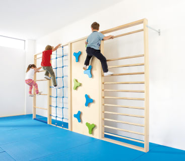 Drei Kinder klettern auf einem Indoor-Holzspielgerät mit Leitern, blauen Seilnetzen und bunten Griffen. Der Boden ist mit blauen Polstern bedeckt, und der Raum ist hell erleuchtet.