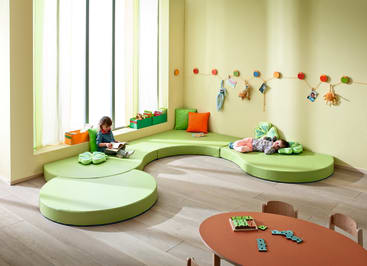 Ein hell erleuchtetes Spielzimmer mit grünen, blattförmigen Sitzkissen, zwei Kindern - eines liest, eines liegt -, einem roten Tisch mit Stühlen und Spielzeug und Dekorationen an einer gelben Wand.