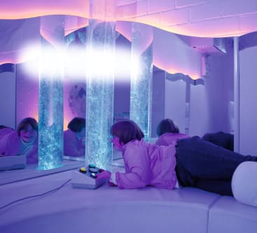 Eine Person liegt auf einem weißen, geschwungenen Sofa in einem Raum mit sanftem violettem Licht und blickt auf ein Paneel mit Knöpfen, in der Nähe von hohen Blasenröhren und Spiegeln, die die beleuchtete Umgebung reflektieren.