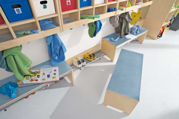 Eine Vorschul- oder Kita-Garderobe mit blauen Bänken, Fächern für Taschen und Kleidung, auf dem Boden aufgereihten Schuhen und einem aufgeschlagenen Bilderbuch auf einer Bank. In den oberen Regalen sind bunte Kisten untergebracht.