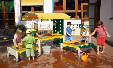 Fünf kleine Kinder spielen mit Wassertischen und Spielzeug in einem Außenbereich mit Fliesenboden. Der Spielbereich ist mit einem gelben Vordach und verschiedenen Behältern ausgestattet. Ein Kind sitzt im Hintergrund an einer Wand.