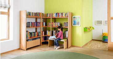 Ein junges Mädchen sitzt auf einer grünen Bank in der Ecke einer hell erleuchteten Kinderbibliothek und liest ein Buch. An den grünen und weißen Wänden stehen Regale mit bunten Büchern. Ein Fenster lässt natürliches Licht herein.
