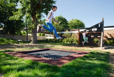 Zwei Kinder spielen auf einem Spielplatz im Freien. Ein Kind befindet sich in der Luft über einem versenkten Trampolin, während ein anderes Kind auf einer Wand mit runden Aussparungen klettert. Der Bereich ist von Gras, Bäumen und einem Gebäude umgeben.
