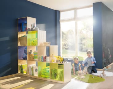 Zwei kleine Kinder spielen mit Bauklötzen auf einem grünen Teppich neben einem modernen Holzregal, das mit Büchern, Spielzeug und Aufbewahrungskörben gefüllt ist. Sonnenlicht strömt durch ein großes Fenster in den hell erleuchteten Raum.