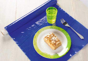 Ein Stück Kuchen mit Puderzucker auf einem Teller mit grünem Rand, eine Gabel und ein Glas mit gelb-grüner Flüssigkeit stehen auf einem glänzend blauen Tischset auf einem hellen Tisch.