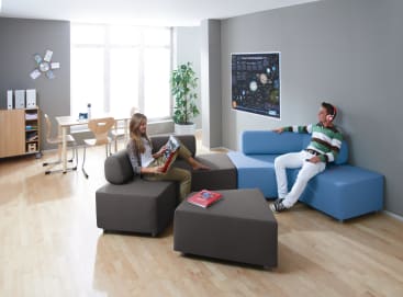 Zwei Personen sitzen auf modernen Sofas in einem hellen Raum mit Holzboden. Eine Person liest eine Zeitschrift, die andere hat Kopfhörer auf. Im Hintergrund stehen ein Tisch, Stühle, Regale und eine Pflanze. An der Wand hängt ein Poster.