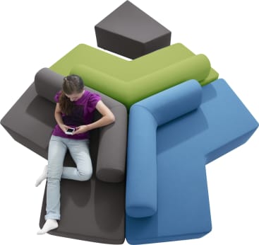 Eine Person in lilafarbenem Hemd und heller Hose sitzt auf einem modularen Sofa mit Teilen in Grau, Grün und Blau und schaut auf ein Smartphone. Ein passender kleiner grauer Hocker steht daneben.