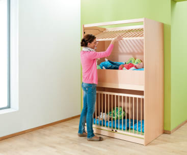 Eine Frau steht neben einem hölzernen Etagenbett. Sie hebt das obere Gitter über einem Baby an, das in der oberen Koje liegt, während ein anderes Baby in der unteren Koje ruht, die ein geschlossenes Tor hat. Der Raum hat grüne und weiße Wände und einen Holzboden.