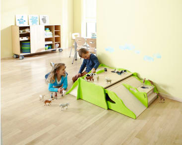 Zwei kleine Kinder spielen auf dem Boden mit Spielzeugtieren, die auf einem Holzspielzeug mit grünen Kanten angeordnet sind, in einem hellen Raum mit hellem Holzboden und Regalen im Hintergrund.