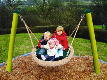 Drei kleine Kinder sitzen zusammen auf einer runden Netzschaukel, die an Seilen und grünen Stangen in einem Park aufgehängt ist. Im Hintergrund sind Bäume und Grasflächen zu sehen.