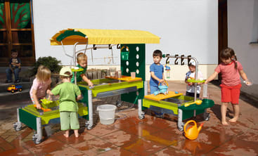 Fünf kleine Kinder spielen draußen auf einer gefliesten Fläche mit einer Wasseraktivitätsstation. Die Station hat grüne und gelbe Teile, Behälter und Gießkannen. Um sie herum wird Wasser auf den Boden gespritzt.