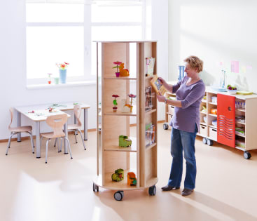 Eine Person steht neben einem hohen, drehbaren Holzregal auf Rädern in einem hellen Klassenzimmer und ordnet Bücher ein. Der Raum hat Tische und Stühle in Kindergröße und Schränke mit Pflanzen und Dekorationen.
