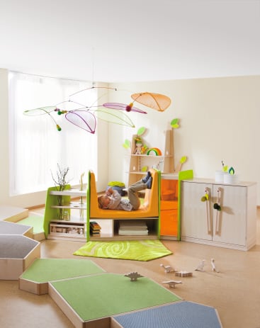 Ein Kind liegt auf einem gelben und orangefarbenen Indoor-Spielgerät in einem hellen, modernen Spielzimmer mit Möbeln aus Naturholz, geometrischen grünen Matten und Dekorationen, die an Blätter erinnern.