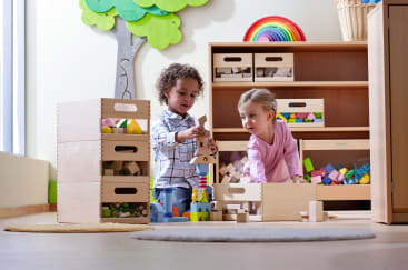 Zwei kleine Kinder spielen mit Holzbausteinen in einem hell erleuchteten Klassenzimmer. In der Nähe sind Holzkisten mit Bauklötzen gestapelt, und an der Wand sind bunte Dekorationen wie ein Regenbogen und ein Baum zu sehen.