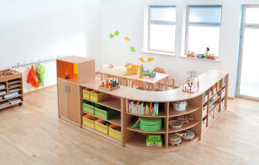 Ein helles Klassenzimmer mit Holzmöbeln, Regalen mit Büchern und Spielzeug, einem Tisch mit kleinen Stühlen, bunten Wanddekorationen und Kindermänteln, die an Haken hängen. Zwei große Fenster lassen natürliches Licht herein.