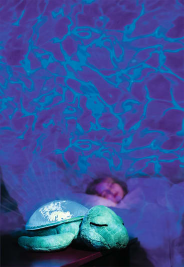 Ein Kind schläft im Bett unter einem violetten und blauen Licht, das wasserähnliche Muster an die Wand projiziert. Im Vordergrund sitzt eine grüne Plüschschildkröte mit einem leuchtenden Panzer auf einer nahen Fläche.