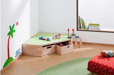Eine Kinderspielecke mit einem Holzregal mit Bauklötzen, einer roten Bank mit einer Puppe, einem grünen Teppich und einer Wand mit einer Palme und Büchern am Fenster.