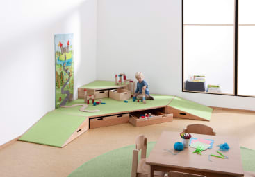 Ein kleines Kind spielt mit Holzklötzen auf einer grünen, gestuften Spielplattform mit Aufbewahrungsschubladen in einem hellen, minimalistischen Raum mit einem Tisch, Stühlen und einem großen Fenster. Eine bunte Spielmatte lehnt an der Wand.
