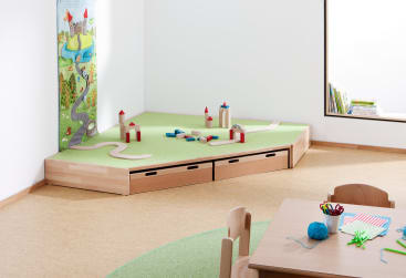 Ein Kinderspielplatz mit einer erhöhten Plattform, Spielzeuggebäuden und Straßen, daneben ein kleiner Tisch mit Stühlen. Im Hintergrund sind ein Fenster und ein Bücherregal mit Büchern zu sehen. Der Raum hat helle Böden und Wände.
