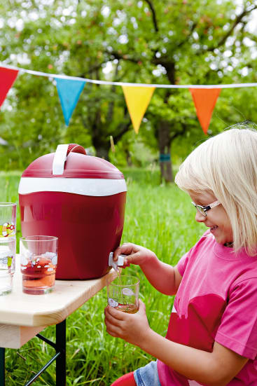 Ein kleines Kind mit Brille und rosa Hemd schenkt ein Getränk aus einer großen roten Kühlbox in ein Glas ein, während es an einem Tisch im Freien sitzt, mit bunten Wimpeln und Grünpflanzen im Hintergrund.