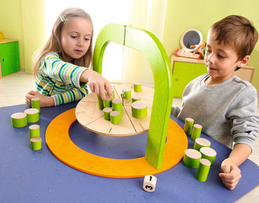 Zwei kleine Kinder sitzen an einem blauen Tisch und spielen ein Balancierspiel mit grünen zylindrischen Figuren auf einer aufgehängten Holzplattform. Ein Würfel mit Zahlen liegt auf dem Tisch. Der Hintergrund ist ein heller, grün gemauerter Raum.