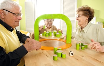 Drei ältere Erwachsene sitzen an einem Tisch und spielen ein Tischstapelspiel mit grünen und beigen zylindrischen Spielsteinen, einem orangefarbenen und grünen Rahmen und einem Würfel mit der Zahl sechs. Sie wirken konzentriert und beschäftigt.