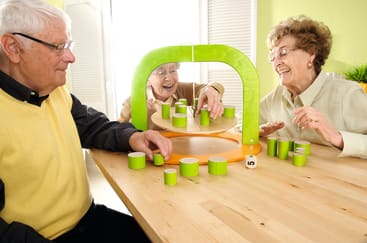 Drei ältere Erwachsene sitzen um einen Tisch herum und spielen ein Tischspiel mit grünen und beigen zylindrischen Spielsteinen und einer rotierenden Plattform. Sie wirken konzentriert und beschäftigt.