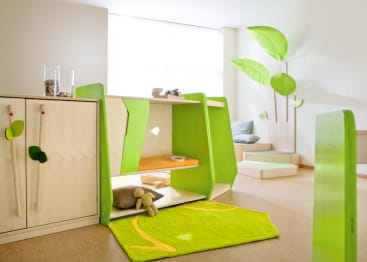 Ein hell erleuchtetes Kinderspielzimmer mit grünen und weißen Möbeln, einem grünen Teppich, einem Plüschtier und einer Wanddekoration mit großen grünen Blättern. Im Hintergrund befindet sich ein Fenster, das natürliches Licht hereinlässt.