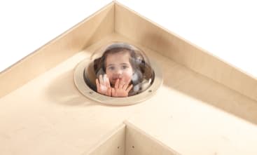Ein kleines Kind schaut durch eine durchsichtige Plastikkuppel, die in die Ecke einer leichten Holzkonstruktion eingebaut ist, und drückt beide Hände gegen die Innenseite der Kuppel.