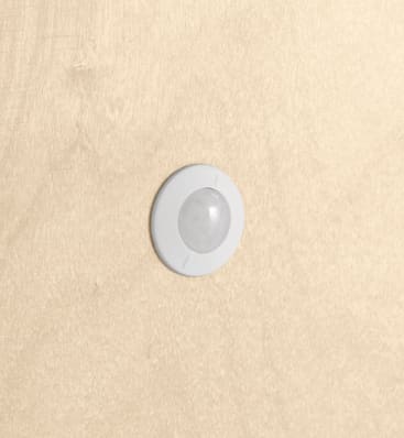 Ein runder, grauer Bewegungssensor ist an einer hellbeigen Wand angebracht. Der Sensor befindet sich in der Mitte des Bildes.