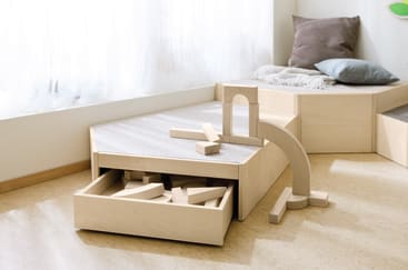 Ein heller Holzspielbereich mit einer Schublade mit Holzbauklötzen, einige Klötze liegen oben und auf dem Boden. Es gibt eine gepolsterte Bank mit Kissen neben einem Fenster, das natürliches Licht hereinlässt.