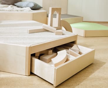 Eine teilweise geöffnete Holzschublade, gefüllt mit verschiedenen Holzbausteinen. Einige Bausteine sind auch auf einer mit Teppich ausgelegten Plattform über der Schublade angeordnet und gestapelt. Der Hintergrund zeigt einen minimalen, hellen Raum.