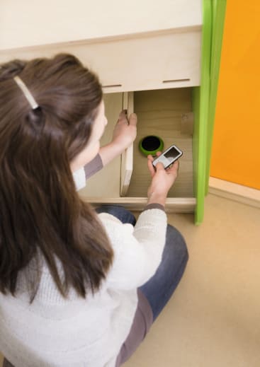 Eine Person mit langen braunen Haaren kniet auf dem Boden und legt ein Smartphone und einen kleinen schwarzen Gegenstand in einen Holzschrank mit grünem Rand.