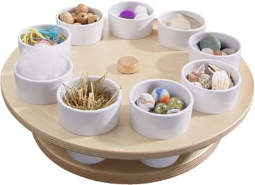 Ein rundes Holztablett enthält acht weiße Schalen mit verschiedenen Gegenständen wie Murmeln, Muscheln, Holzperlen, Stroh, Baumwolle, Schnur, Sand und Pompons. In der Mitte des Tabletts befindet sich ein Holzknauf.