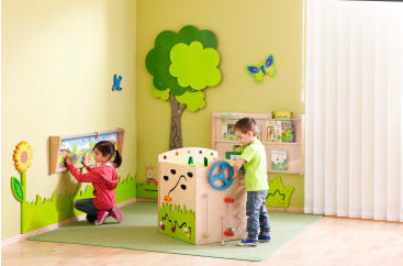 Zwei kleine Kinder spielen in einer hell erleuchteten Ecke eines Klassenzimmers mit grünen Wänden. Eines nutzt ein an der Wand befestigtes Aktivitätsbrett, während das andere ein Rad auf einer hölzernen Spielstruktur dreht. Der Raum ist mit Motiven zum Thema Natur dekoriert.