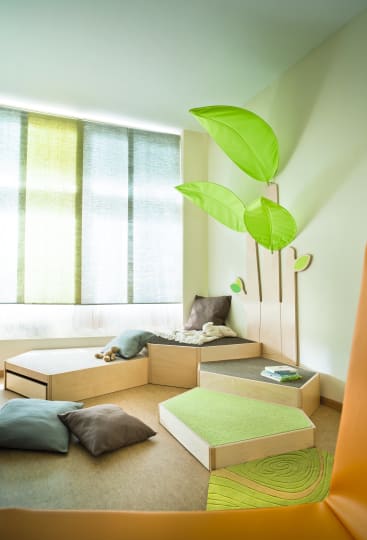 Ein helles Spielzimmer mit Sonnenlicht, das durch ein großes Fenster fällt, gepolsterten Sitzblöcken, grünen Blattdekorationen an der Wand, verstreuten Kissen und Kinderspielzeug und Büchern auf den Podesten.