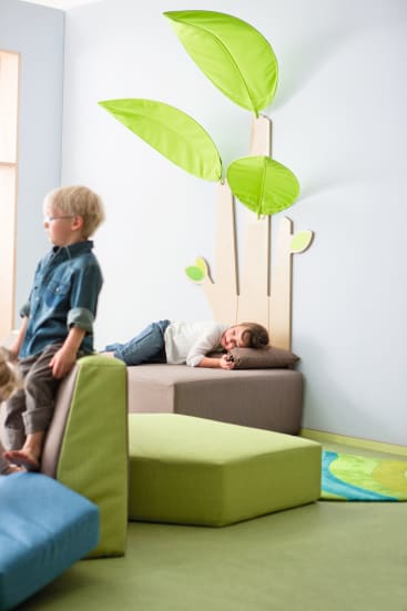 Ein Kind liegt auf einer gepolsterten Bank unter dekorativen grünen Blattformen, die an der Wand befestigt sind, während ein anderes Kind im Vordergrund auf Schaumstoffspielblöcken in einem hellen Spielzimmer steht.