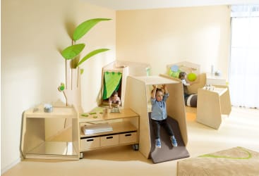 Drei kleine Kinder spielen in einem lichtdurchfluteten Raum mit hellgelben Wänden, hölzernen Spielmöbeln und grünem Blattschmuck. Ein Kind hängt an einer Stange, während die anderen an verschiedenen Spielstationen sitzen.