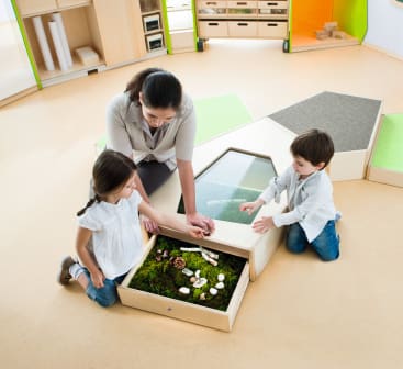 Ein Erwachsener und zwei Kinder untersuchen ein Tablett mit Moos, Pilzen und Tannenzapfen in einem hellen Klassenzimmer mit Holzmöbeln und Regalen.