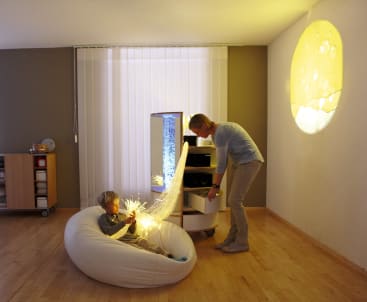 Ein Kind sitzt auf einem weißen Sitzsack und hält faseroptische Stränge, die Licht aussenden, während ein Erwachsener in der Nähe steht und in einen Schrank schaut. Ein gelbes Licht, das dem Mond ähnelt, wird in einem schwach beleuchteten Raum an die Wand projiziert.