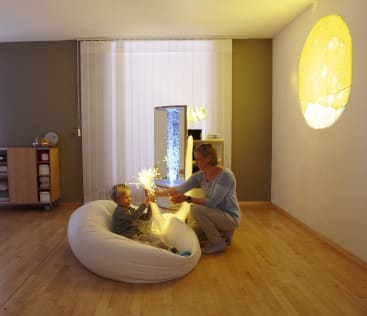 Ein Erwachsener und ein Kind interagieren mit faseroptischen Lichtern in einem sanft beleuchteten Sinnesraum. Das Kind sitzt auf einem weißen Sitzsack, während der Erwachsene in der Nähe kniet. Ein gelbes Lichtmuster wird an die Wand projiziert.