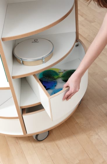 Eine Hand zieht eine Schublade in einem weißen, runden Aufbewahrungsschrank auf Rädern auf und enthüllt bunte, transparente Scheiben. Ein Tamburin liegt auf einem Regal über der geöffneten Schublade. Der Boden ist aus hellem Holz.