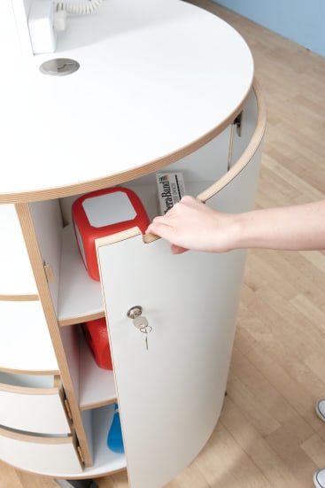 Die Hand einer Person öffnet mit einem Schlüssel eine gewölbte, weiße Schranktür. Im Inneren des Schranks befinden sich ein rot-weißer Erste-Hilfe-Kasten und eine Zeitung in einem Holzregal. Der Schrank steht auf einem hellen Holzboden.