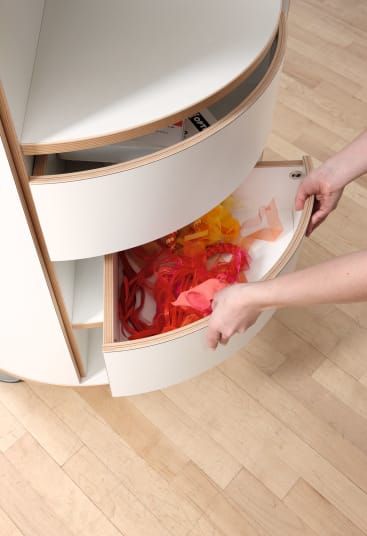 Eine Person öffnet eine gebogene Schublade in einem weißen, runden Schrank. In der Schublade befinden sich rote, orangefarbene und gelbe durchsichtige Stoffstreifen. Der Schrank steht auf einem hellen Holzboden.