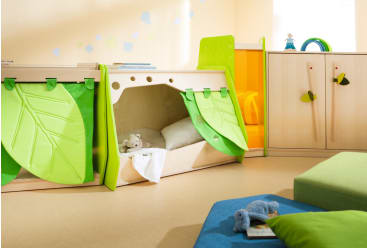 Ein Spielzimmer für Kinder mit einer hölzernen Schlafecke mit grünen blattförmigen Vorhängen, einem Kissen und einem Stofftier darin. Außerdem gibt es einen Holzschrank und weiche Spielmatten auf dem Boden.