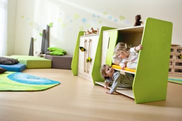 Zwei kleine Kinder spielen in einem grünen und holzfarbenen Spielgerät in einem hell erleuchteten Raum mit weichem Bodenbelag, Kissen und einfachen Dekorationen. Ein Kind befindet sich im unteren Teil und das andere im oberen Teil.