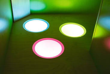 Drei kreisförmige Lichter - ein weißes, ein grünes und ein rosafarbenes - sind in eine hölzerne Oberfläche eingelassen und beleuchten die umliegenden Wände in ihren jeweiligen Farben.